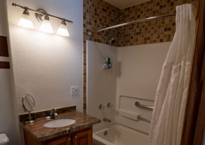 A bathroom in this lovely vacation home features a bathtub and shower, tiled backsplash, a sink with a mirror, overhead lighting, and a shower curtain. There are mounted soap dispensers and wooden cabinets under the sink—perfect for your stay with Alpenglow Vacation Rentals.