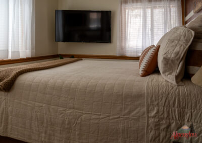 A neatly made bed with beige linens and a patterned pillow sits under two windows in a cozy bedroom with a wall-mounted TV, perfect for enjoying your stay at our vacation home rentals.