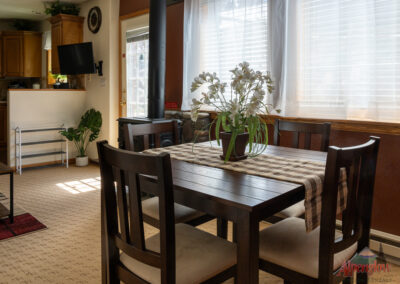 A dining area features a dark wooden table with a checkered runner and a potted plant centerpiece. Adjacent is a kitchen with wooden cabinets, a TV, plants, and sunlight streaming through the windows. This inviting space is perfect for those seeking cozy vacation home rentals.