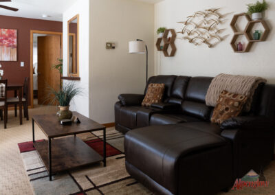 A cozy living room with a brown leather sectional sofa, a wooden coffee table, wall decor featuring metallic bird sculptures, and plants. An open doorway leads to the dining area and other rooms, offering a welcoming retreat perfect for short term house rentals at Alpenglow Vacation Rentals.
