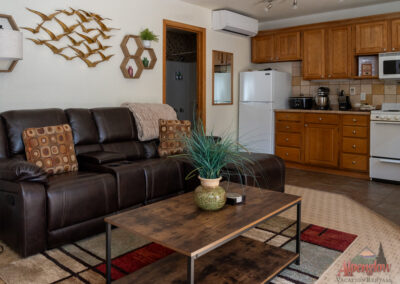A cozy living room in this vacation home features a brown leather couch, wooden coffee table, and an adjoining kitchen area with wooden cabinets, white appliances, and various kitchen items. The décor includes charming wall art and a small plant, making it perfect for vacation rentals.