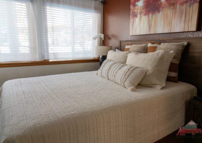 A neatly made bed with white textured linens, multiple pillows, and a wooden headboard invites you to relax. A large window with white curtains is in the background. On the wall, a painting and an Alpenglow Vacation Rentals logo hint at your dreamy vacation home.