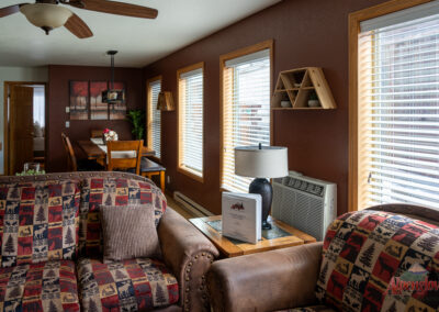 A cozy living room in our vacation home rentals features patterned armchairs, wooden blinds on the windows, a dining table with chairs, a table lamp, and an air conditioning unit mounted on the wall.
