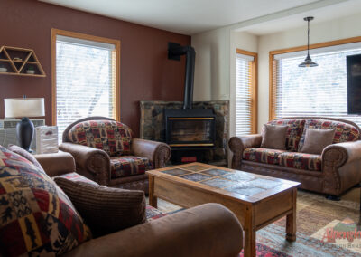 Living room with two patterned sofas, a patterned armchair, a wooden coffee table, and a stone fireplace beneath a TV. Windows with blinds on three sides. Air conditioning unit in the corner. Perfect for relaxing after days exploring; discover more at Alpenglow Vacation Rentals.