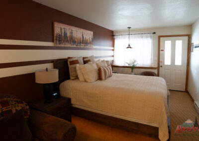 A cozy bedroom with a large bed, numerous pillows, a nightstand with a lamp, a small wall painting, a window with blinds and sheer curtains, an armchair, and a door to the outside—perfect for your stay at our Alpenglow Vacation Rentals.