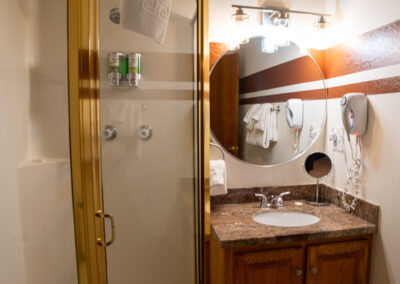 A bathroom in this Alpenglow vacation rental features a glass shower enclosure, a vanity with a granite countertop, a round mirror, and wall-mounted toiletries. The space includes modern lighting and a hairdryer on the wall—perfect for your short term house rental needs.