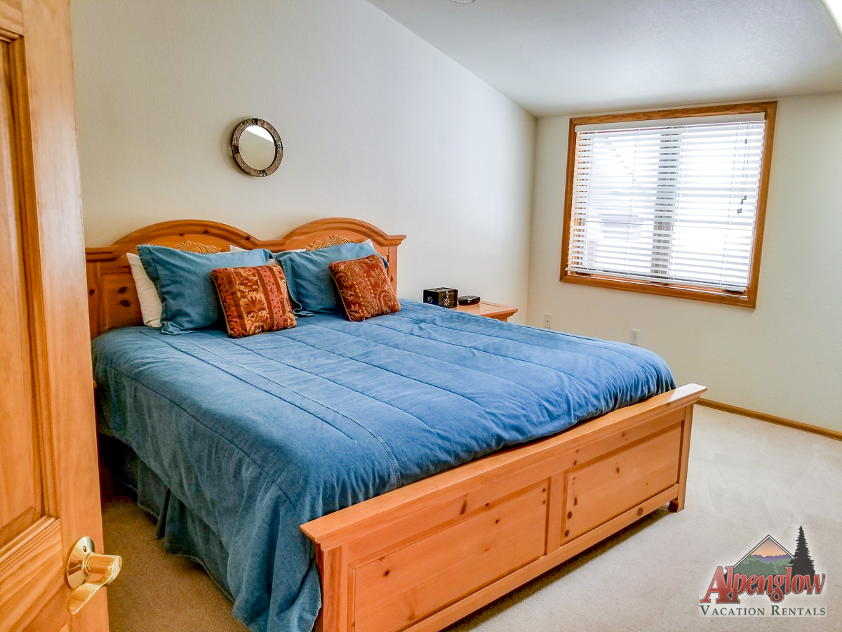 Bedroom with a large wooden bed, blue bedding, two pillows, and a small round mirror on the wall. A window with blinds is on the right. A logo for Alpenglow Vacation Rentals is in the corner.