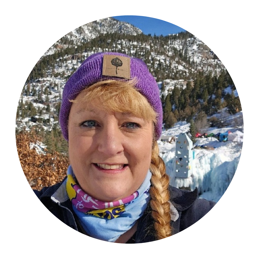 A person with a braided hairstyle wears a purple beanie and neck gaiter, smiling in front of snowy mountains and trees.