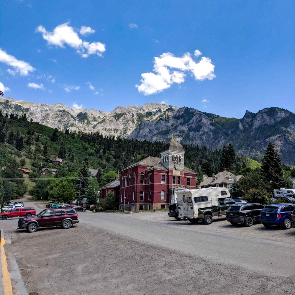 A small town scene with parked cars, a vintage red building with a clock tower, and mountains in the background under a blue sky.