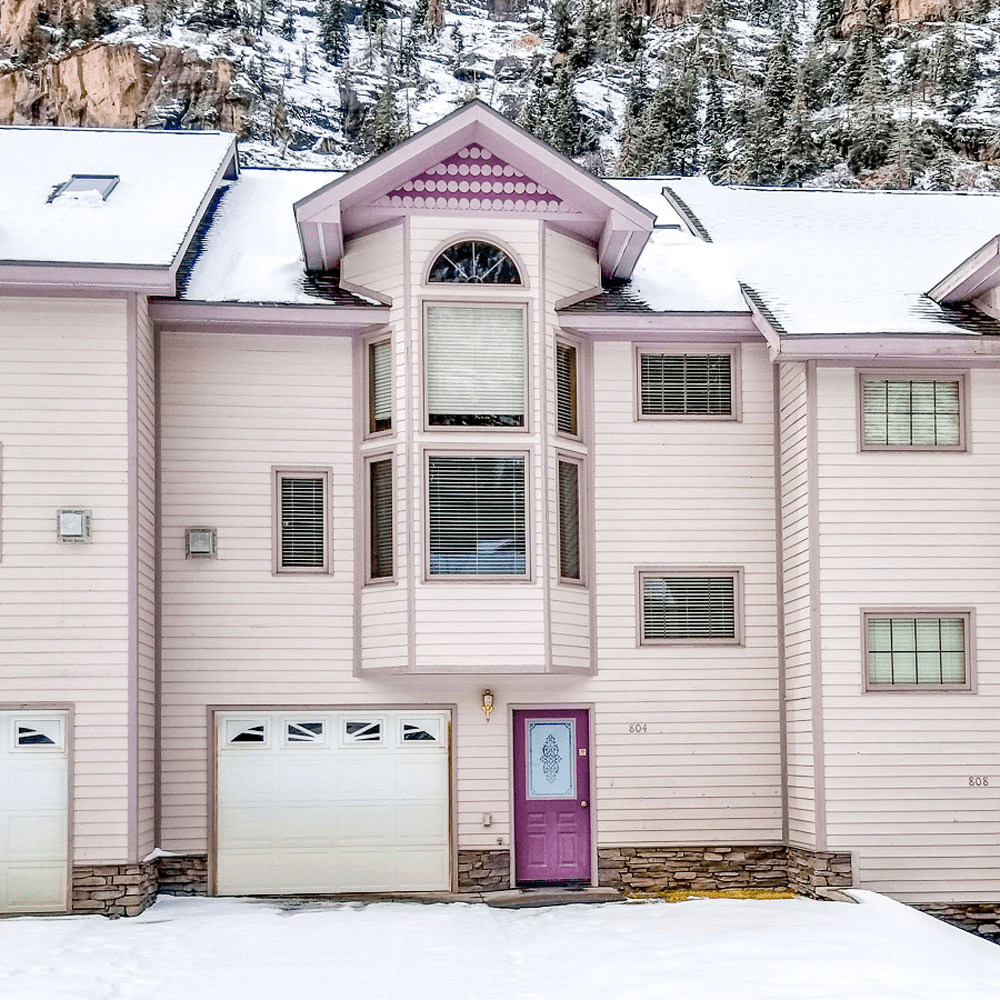 Purple-trimmed townhouse with snowy exterior, featuring bay windows and a garage, set against a rocky, snow-dusted hillside.