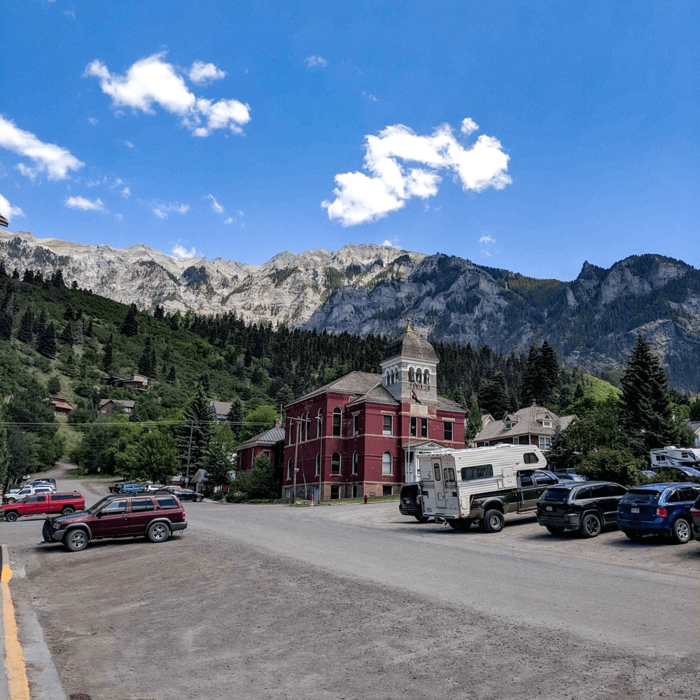 A red building with a tower stands against a mountainous backdrop. Cars are parked along the dirt road under a clear blue sky with scattered clouds.