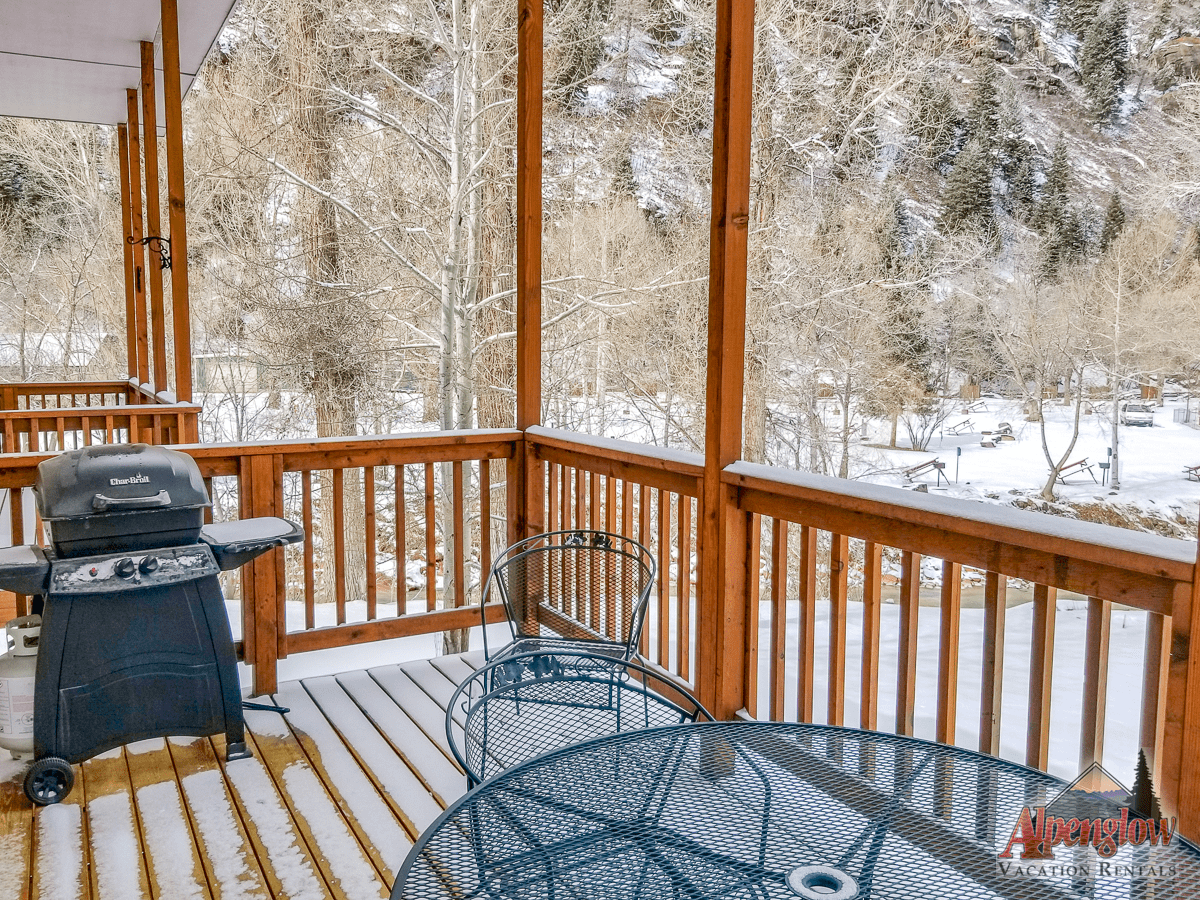 Snow-covered deck with a grill and metal furniture overlooking a wintry wooded landscape.