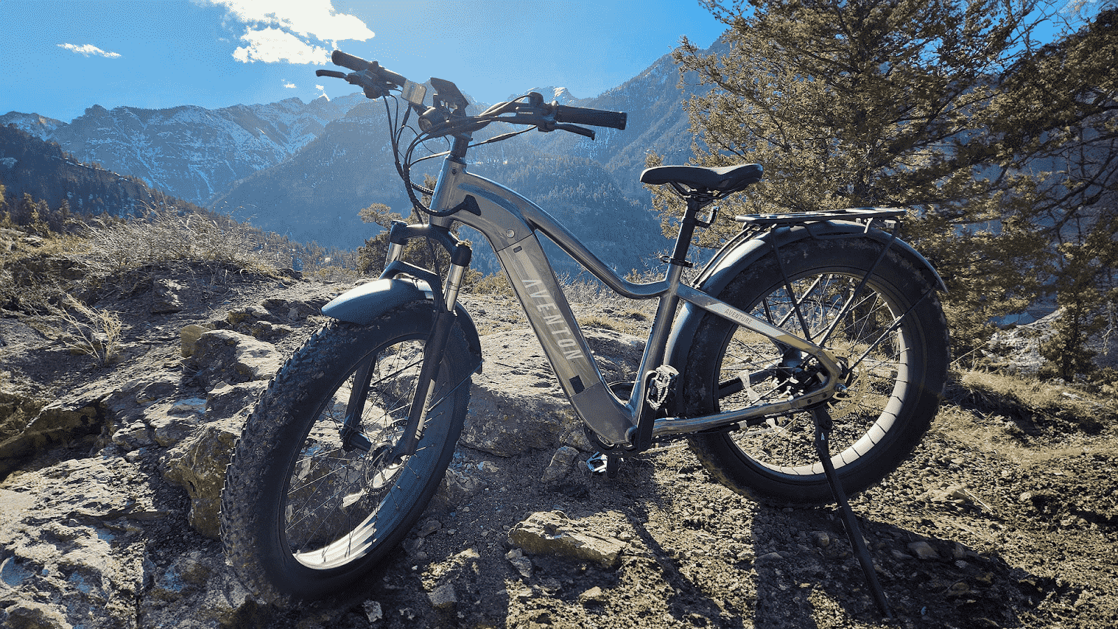 A fat-tire electric mountain bike is parked on a rocky trail with mountains and trees in the background under a blue sky.