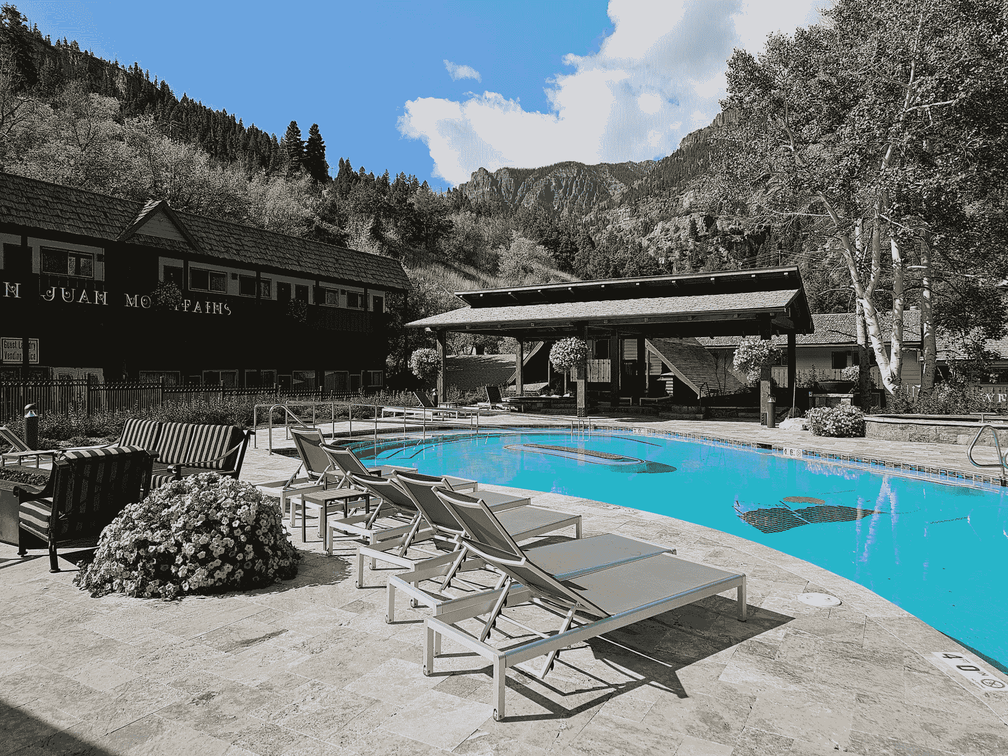 Outdoor swimming pool area with lounge chairs, a covered patio, and a building in the background, set against a backdrop of trees and mountains.