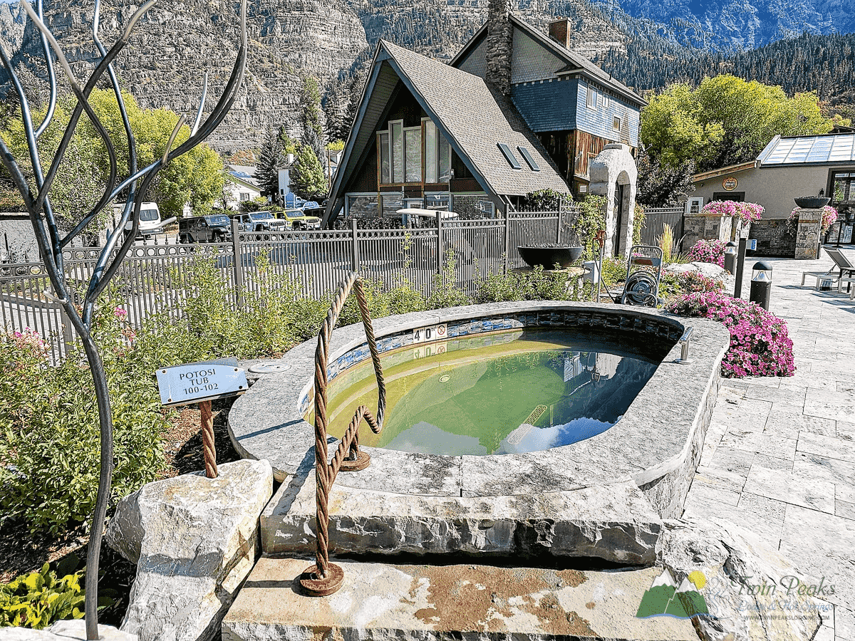 Outdoor oval-shaped stone hot tub with greenish water, surrounded by flowers and fencing, set in a mountain village with houses and trees in the background.