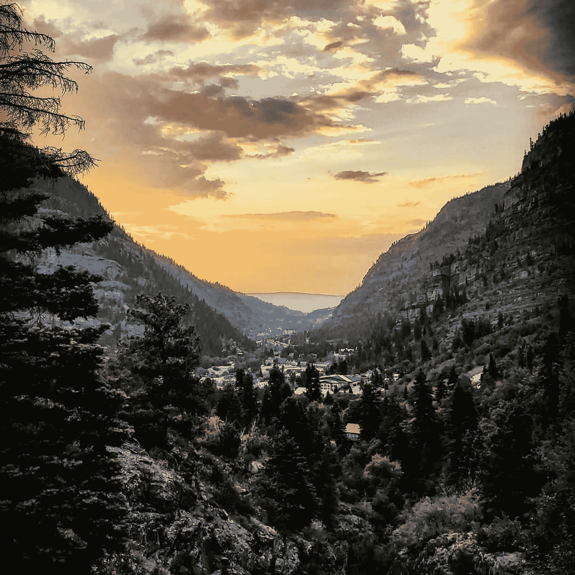 A mountain valley at sunset with a town nestled between forested hills, under a partly cloudy sky with golden light.