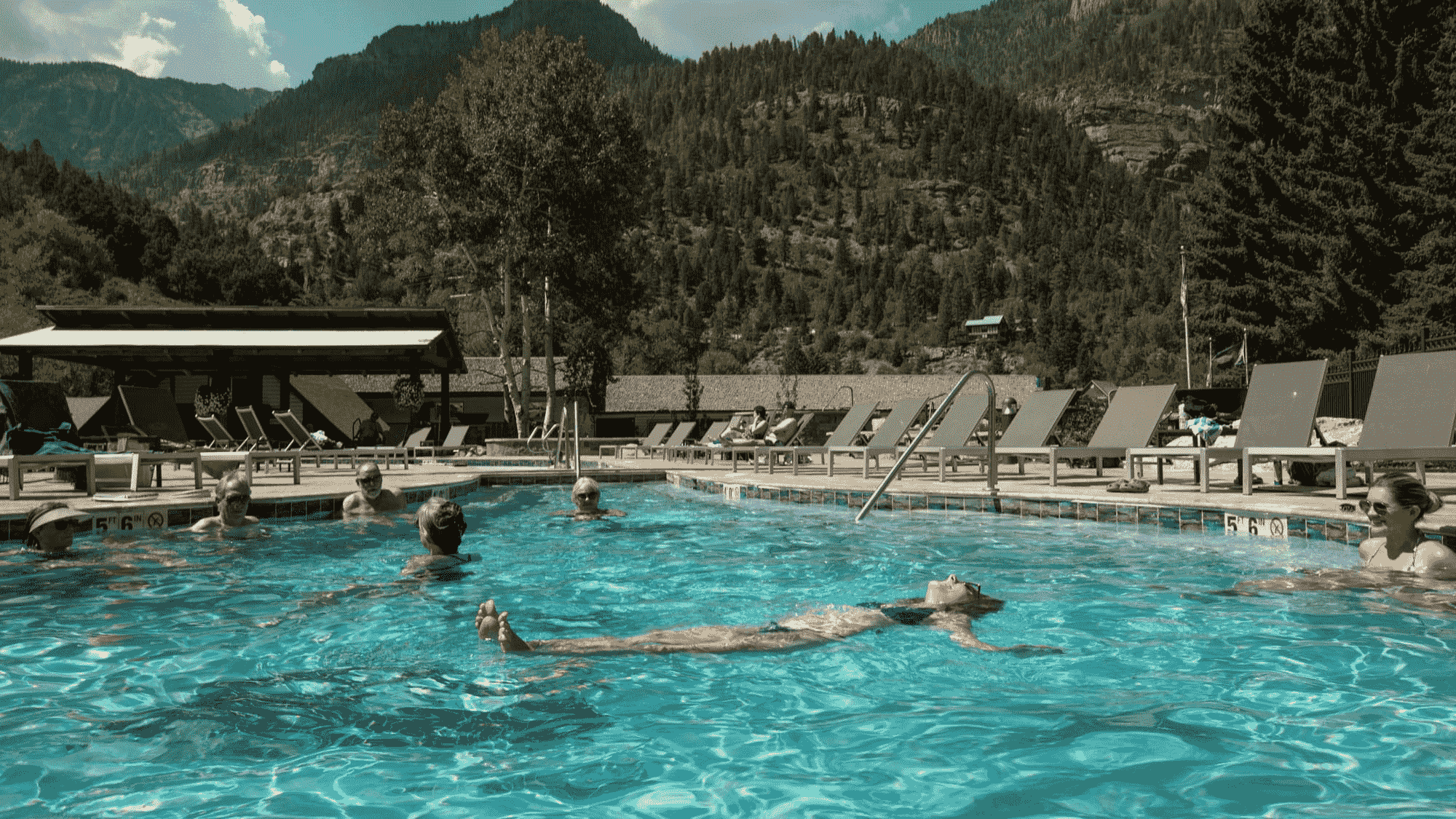 People are relaxing in an outdoor swimming pool surrounded by lounge chairs and mountains under a partly cloudy sky.