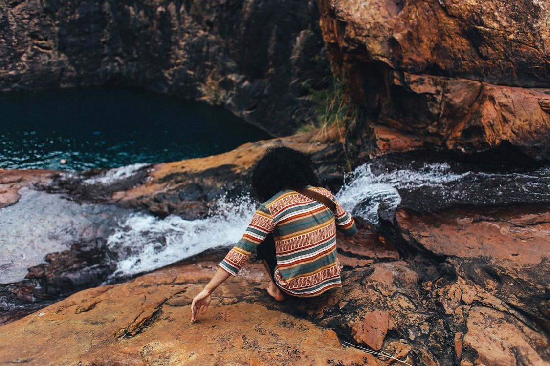 Person wearing a patterned shirt sits on rocky terrain beside a small stream of water flowing toward a blue pool.