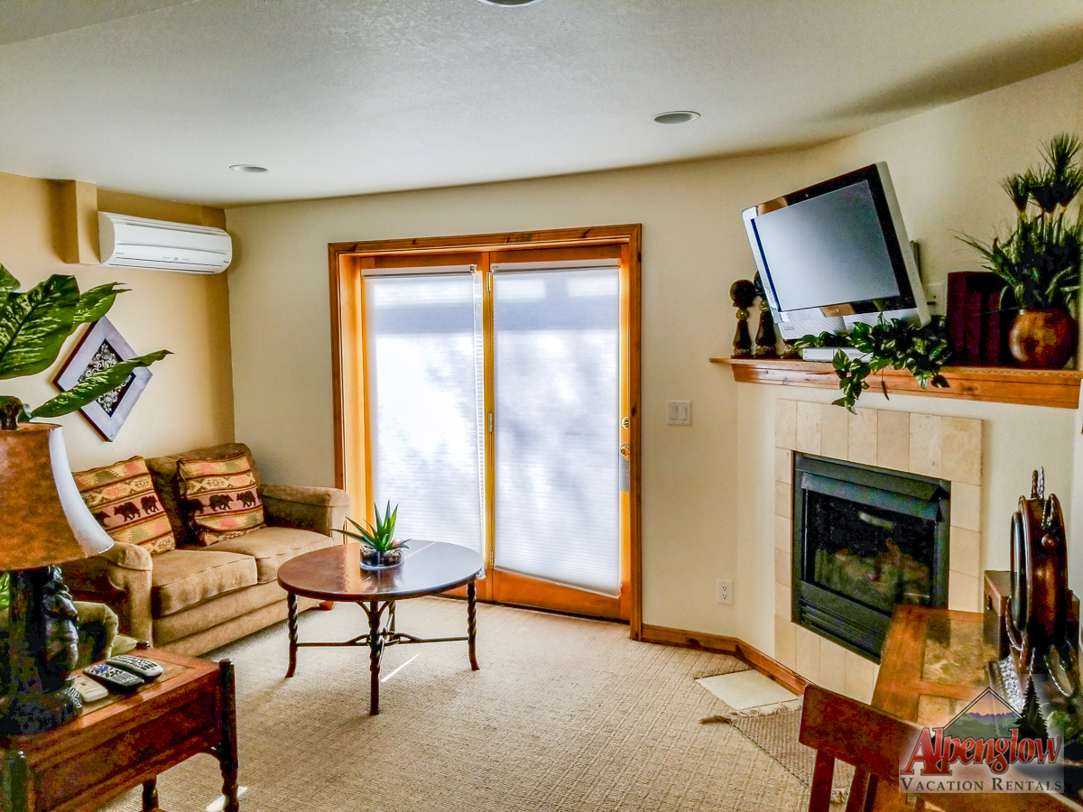 A living room with a sofa, coffee table, fireplace, wall-mounted TV, and large glass doors letting in natural light.