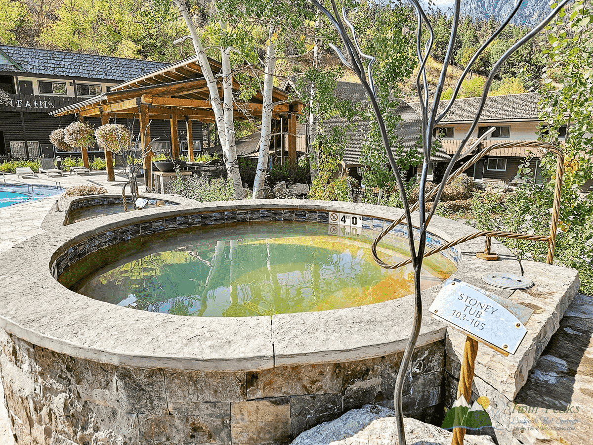 Circular stone hot tub labeled "Stoney Tub 102–105°F" outdoors near buildings, with trees and mountains in the background.
