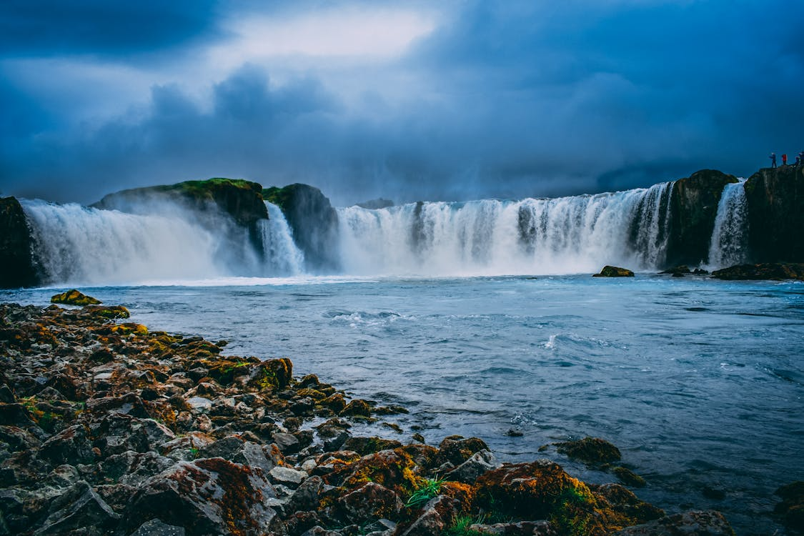 Wide waterfall cascades over rocky cliffs into a river under a cloudy sky, with moss-covered rocks in the foreground.