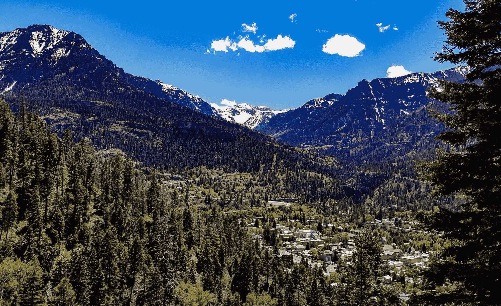 Mountain landscape with dense evergreen forests, a small town nestled in the valley, and snow-capped peaks under a bright blue sky with scattered clouds.