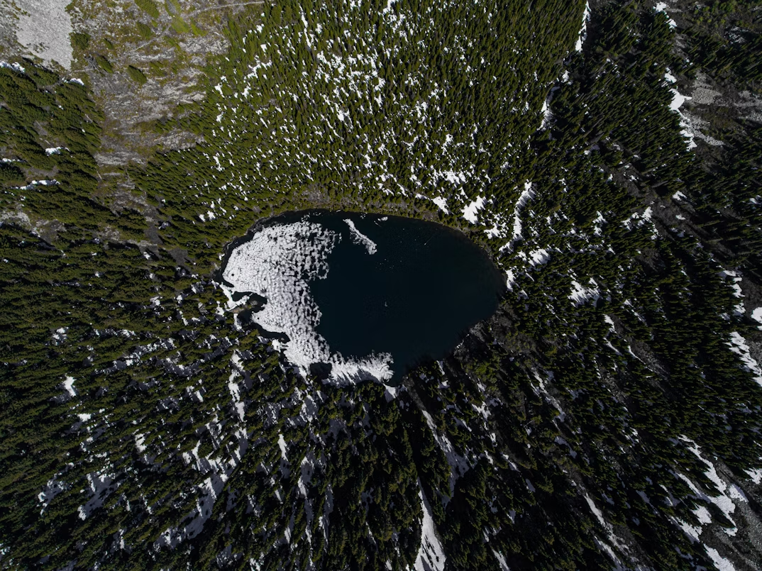 Aerial view of a small, oval-shaped forest lake with snow patches along its edge, surrounded by dense green trees.