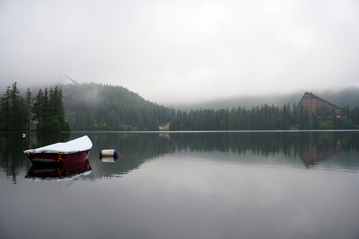 A red boat covered with a tarp floats on a calm lake surrounded by trees and mist, with a triangular building visible in the distance.