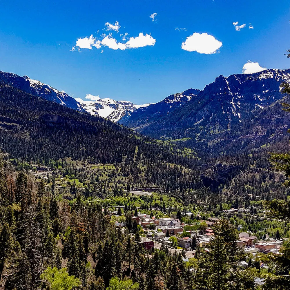 Mountain landscape with dense evergreen forests, a small town nestled in the valley, and snow-capped peaks under a bright blue sky with scattered clouds.