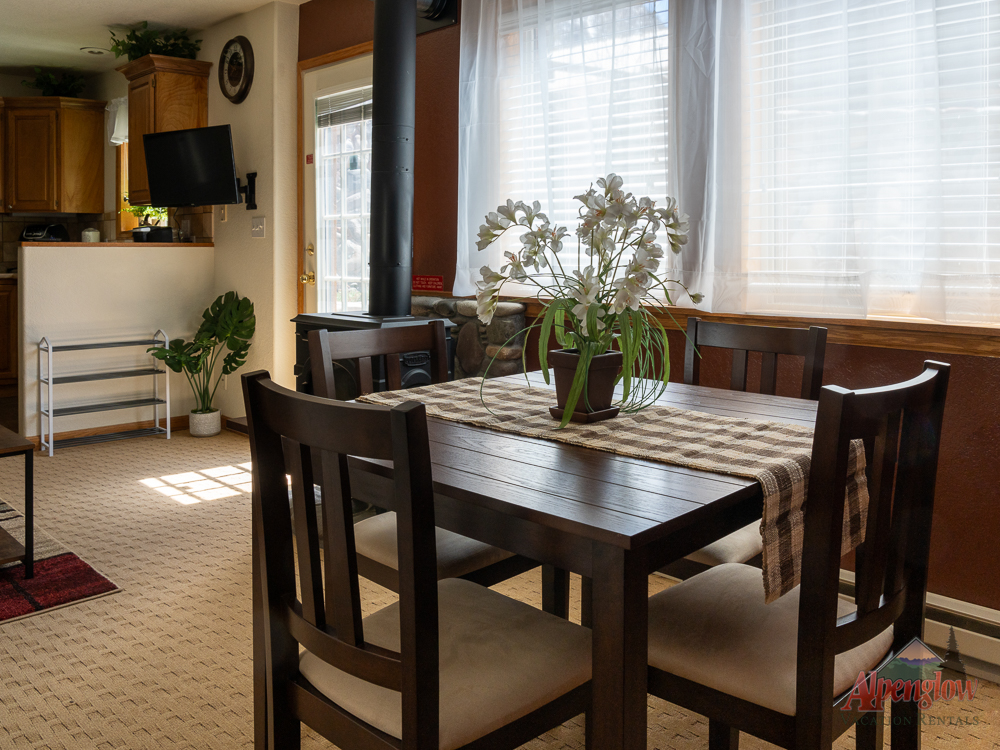 A dining table with four chairs and a checkered runner, topped with a potted plant, sits in a sunlit room next to large windows and a kitchen area in the background.