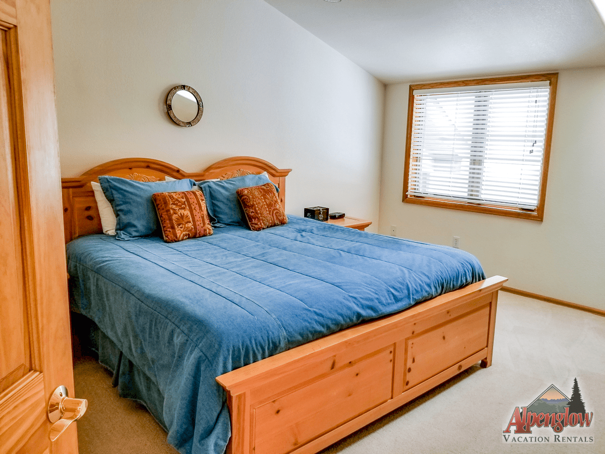 Bedroom with a large wooden bed frame, blue bedding, two orange accent pillows, a small round wall mirror, window with blinds, and beige carpet. Alpenglow Vacation Rentals logo in corner.