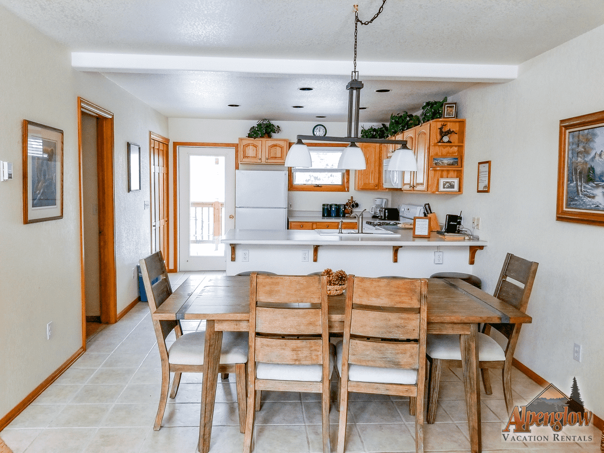 A cozy kitchen and dining area with wooden table and chairs, white countertops, hanging lights, and light-colored cabinets. "Alpenglow Vacation Rentals" logo in the bottom right corner.