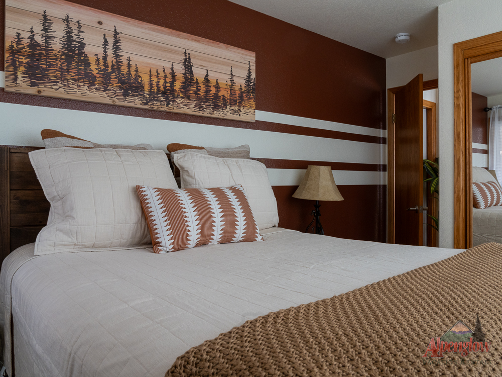 A neatly made bed with beige and brown bedding, a patterned pillow, a wooden headboard, and a forest-themed wall art in a bedroom with brown and white walls.