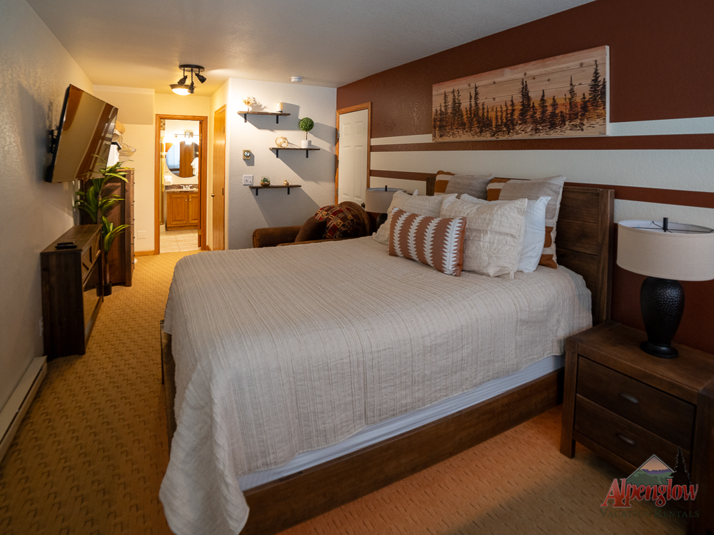 A cozy hotel room with a king bed, neutral bedding, nightstands, a mounted TV, and wall art. A bathroom is visible through an open door at the far end.