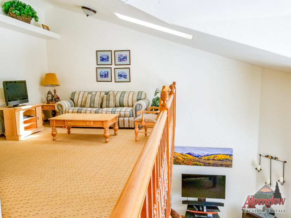 A loft living area with a striped sofa, wooden coffee table, TV on a stand, and framed art on white walls, overlooking a lower level with another TV.
