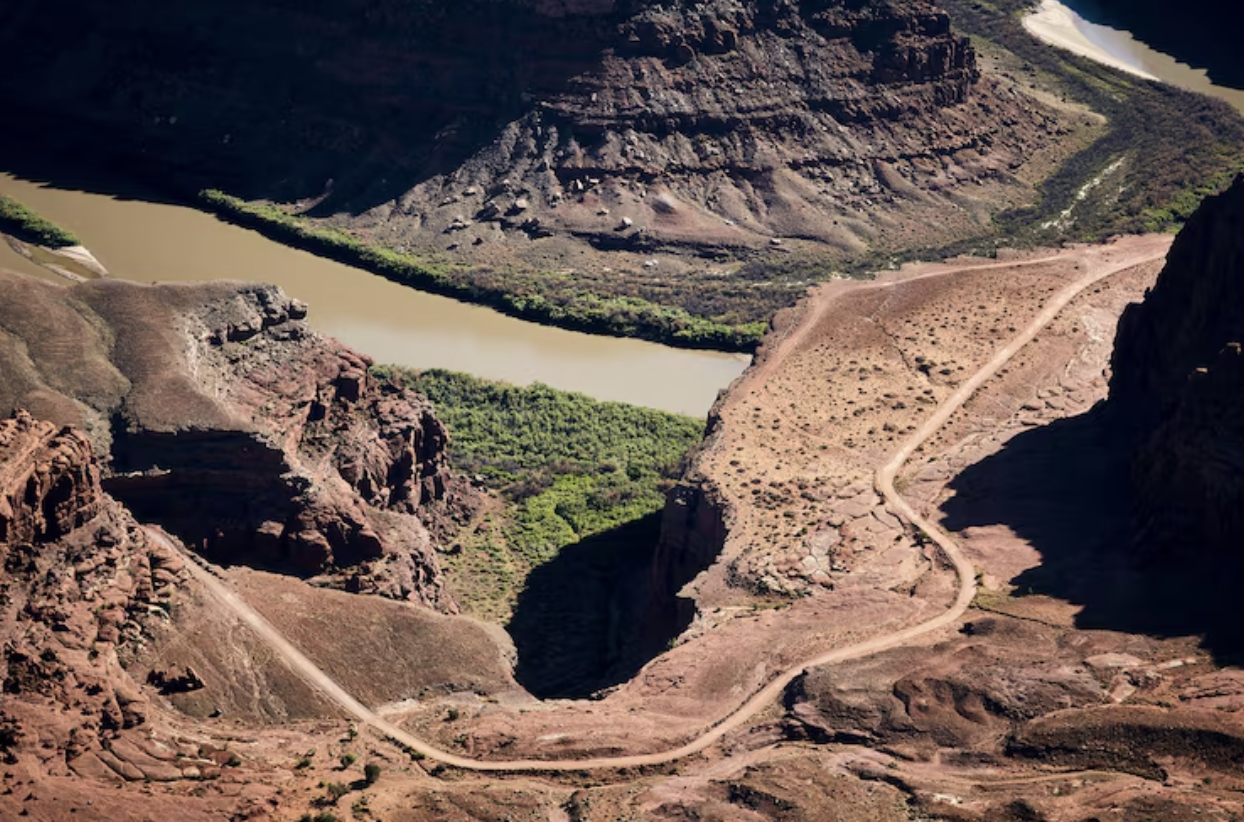 A winding dirt road runs through a rocky desert canyon landscape beside a brown river lined with green vegetation.