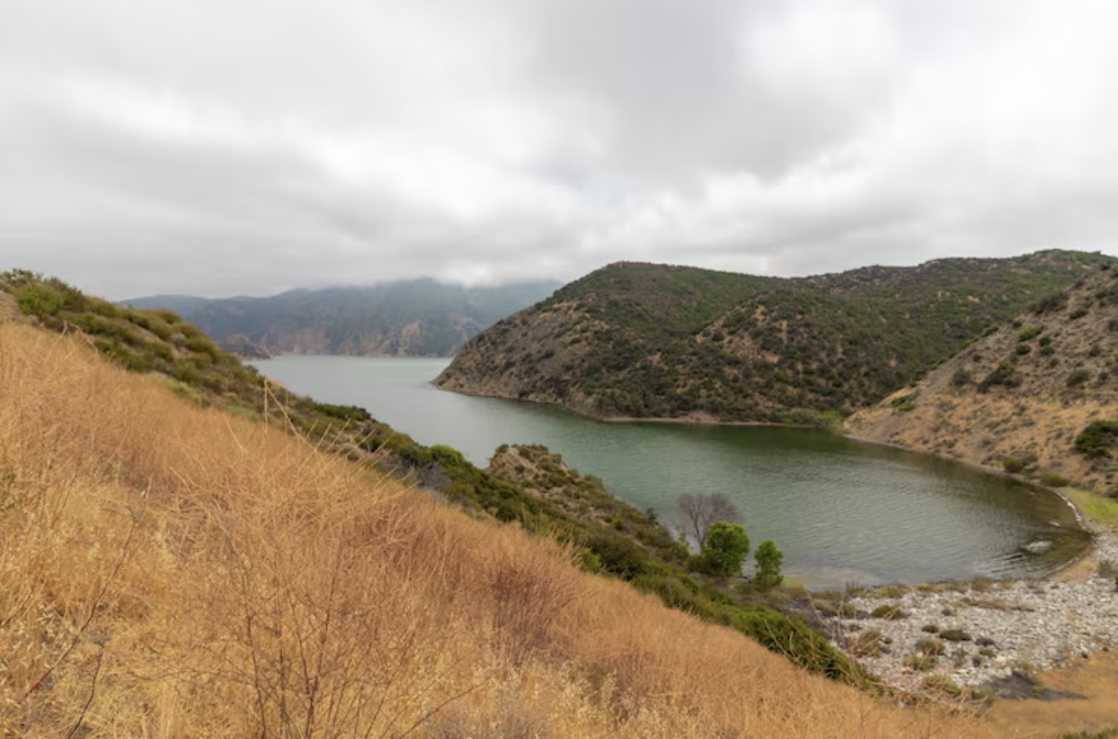 A cloudy sky over a lake surrounded by hills, with dry grass and sparse vegetation in the foreground.