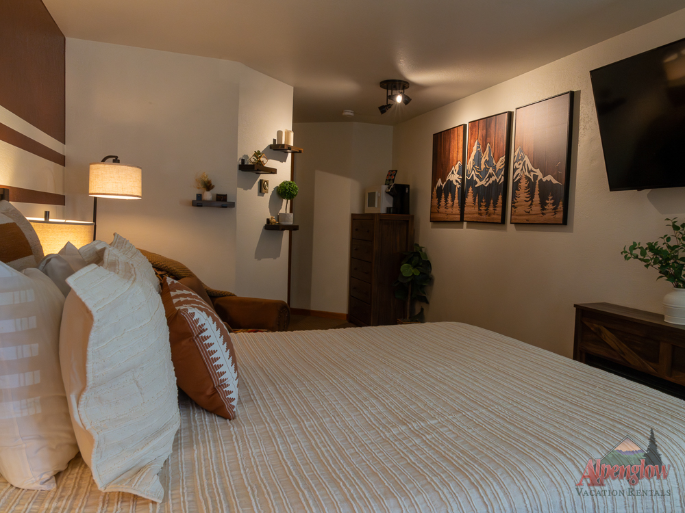 A cozy bedroom with a neatly made bed, decorative pillows, wall-mounted shelves, dresser, TV, and mountain artwork on the wall. The room has warm lighting and neutral tones.