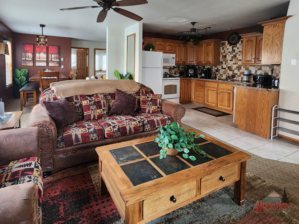 Open-plan living room and kitchen with rustic wooden furniture, patterned sofa, coffee table, and a kitchen area with wood cabinets and appliances.