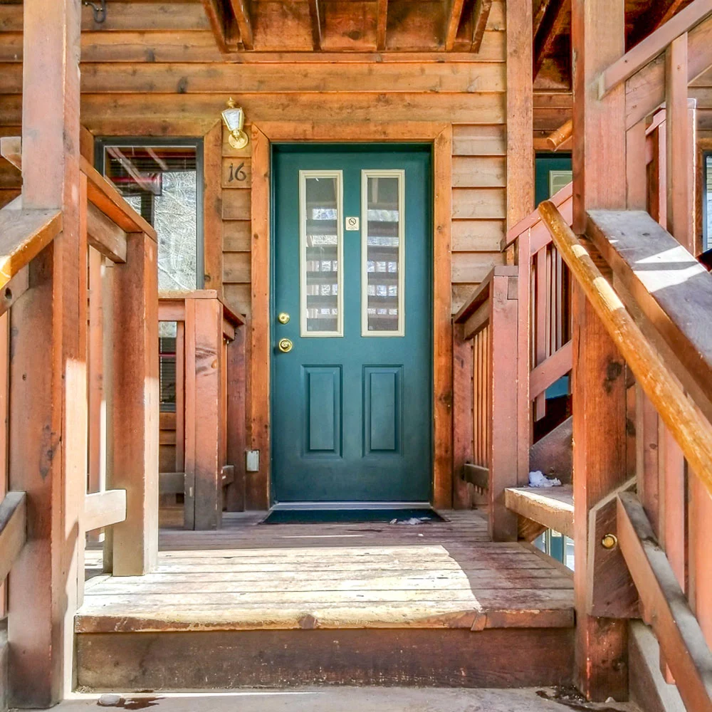 A green door with glass panels, numbered 16, is flanked by wooden railings and a porch in a log cabin-style building, embodying the charm of Alpenglow Vacation Rentals.