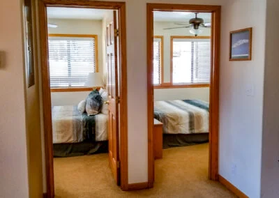 A hallway with two open doorways leading to bedrooms, each featuring a bed with striped bedding. The Alpenglow Vacation Rentals logo is visible in the bottom right corner.